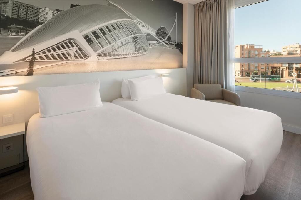 Habitació d'hotel amb dos llits, una finestra gran amb vista a la ciutat i un mural modern a la paret.