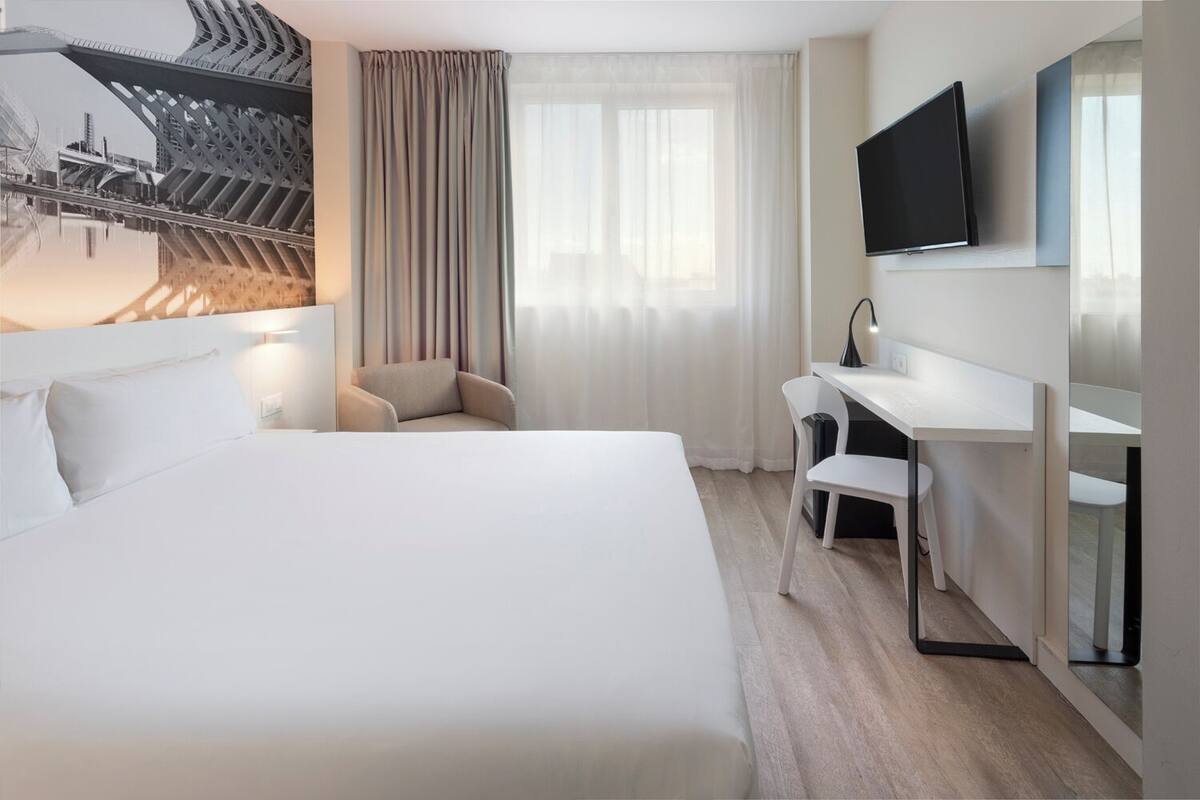 Une chambre d’hôtel moderne dotée d’un grand lit, d’un bureau avec une chaise, d’une télévision à écran plat et offrant une vue sur un pont.