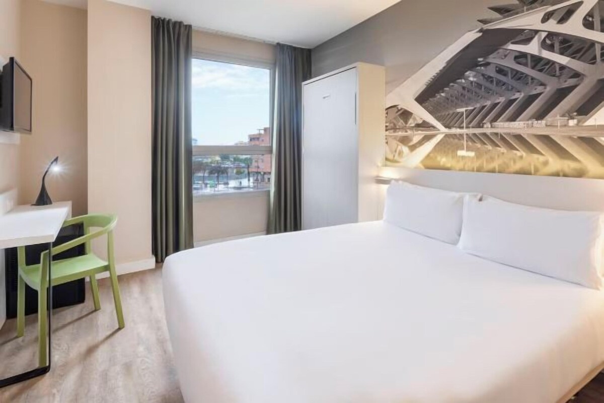 A modern hotel room with a large bed, a desk with a chair, and a view of the city through the window.