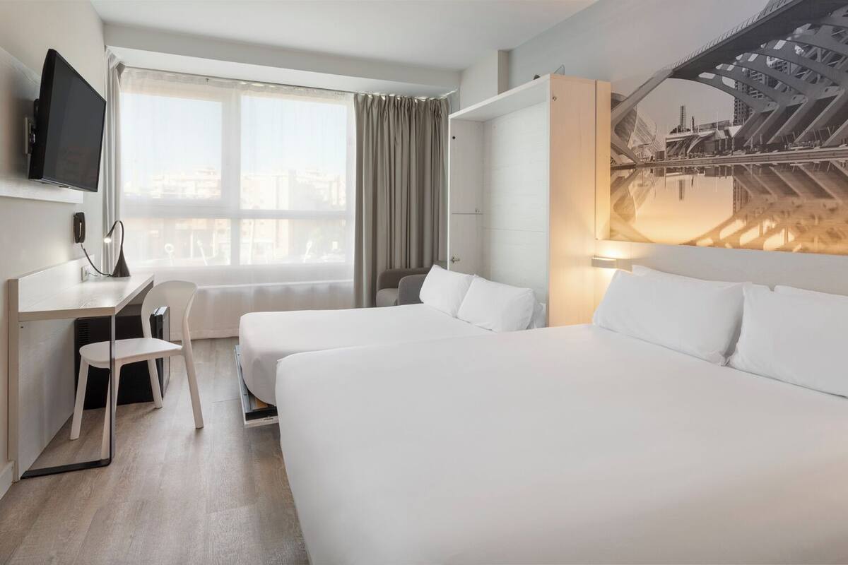 Une chambre d’hôtel avec deux lits, un bureau avec une chaise, une télévision et une grande fenêtre avec des rideaux.