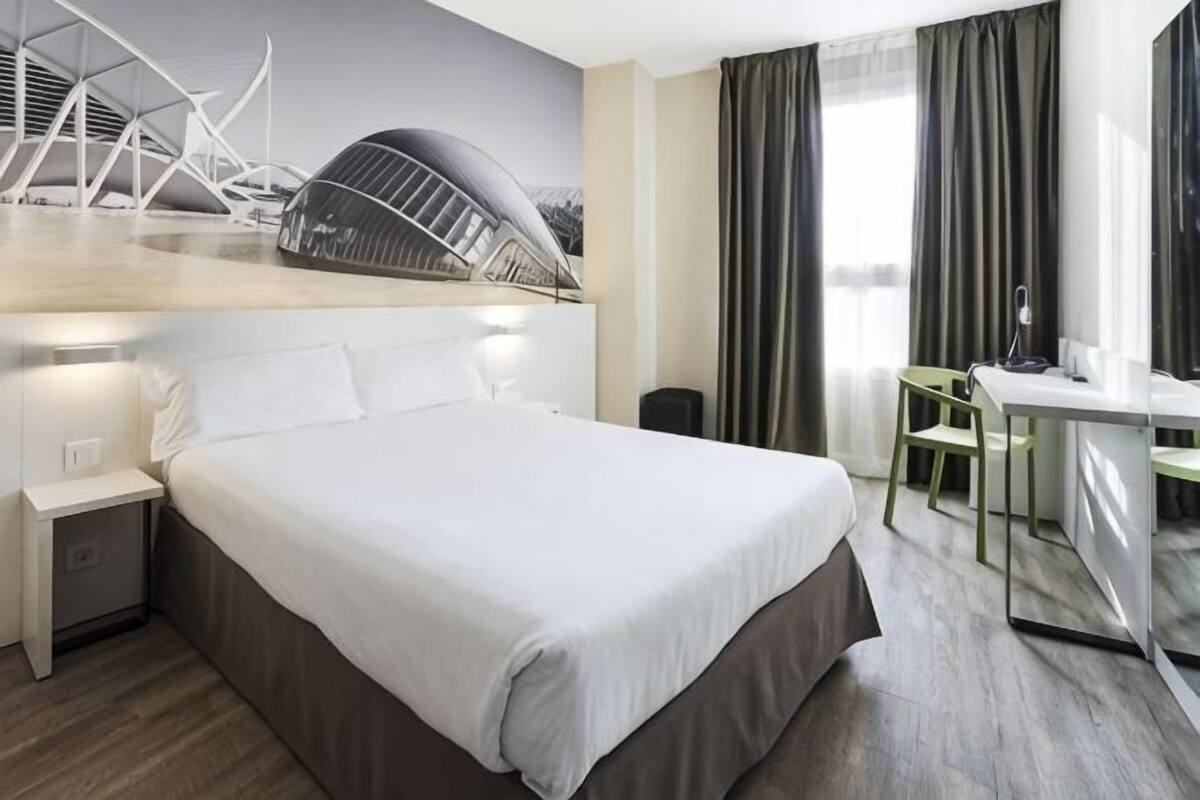 A modern hotel room with a large bed, a desk, and a view of a bridge.