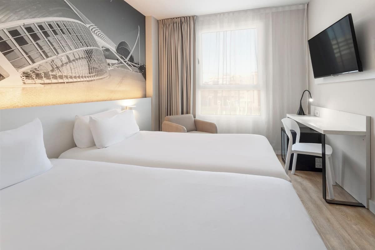 Une chambre d’hôtel avec deux lits, un bureau, une chaise, une télévision et une grande fenêtre avec des rideaux.