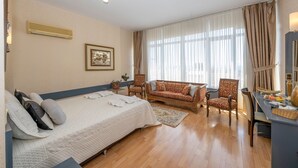 Quarto de hotel com cama, sofá, poltrona, escrivaninha e uma janela grande com cortinas.