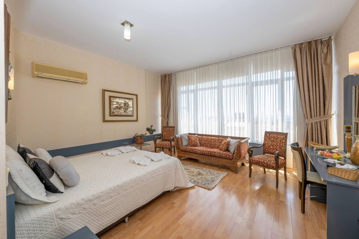 A hotel room with a bed, a sofa, a chair, a desk, and a large window with curtains.