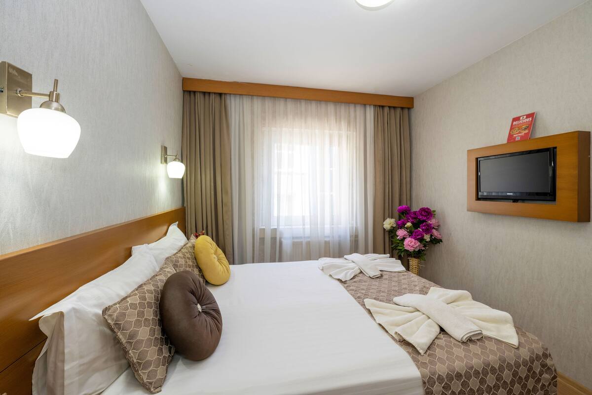 A hotel room with a large bed, two pillows, a TV mounted on the wall, and a window with curtains.