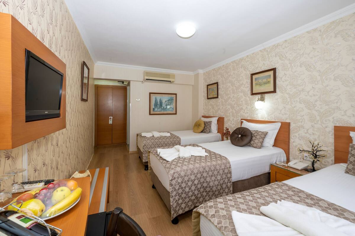 A hotel room with three beds, a flat-screen TV, a fruit bowl, and a desk with a chair.