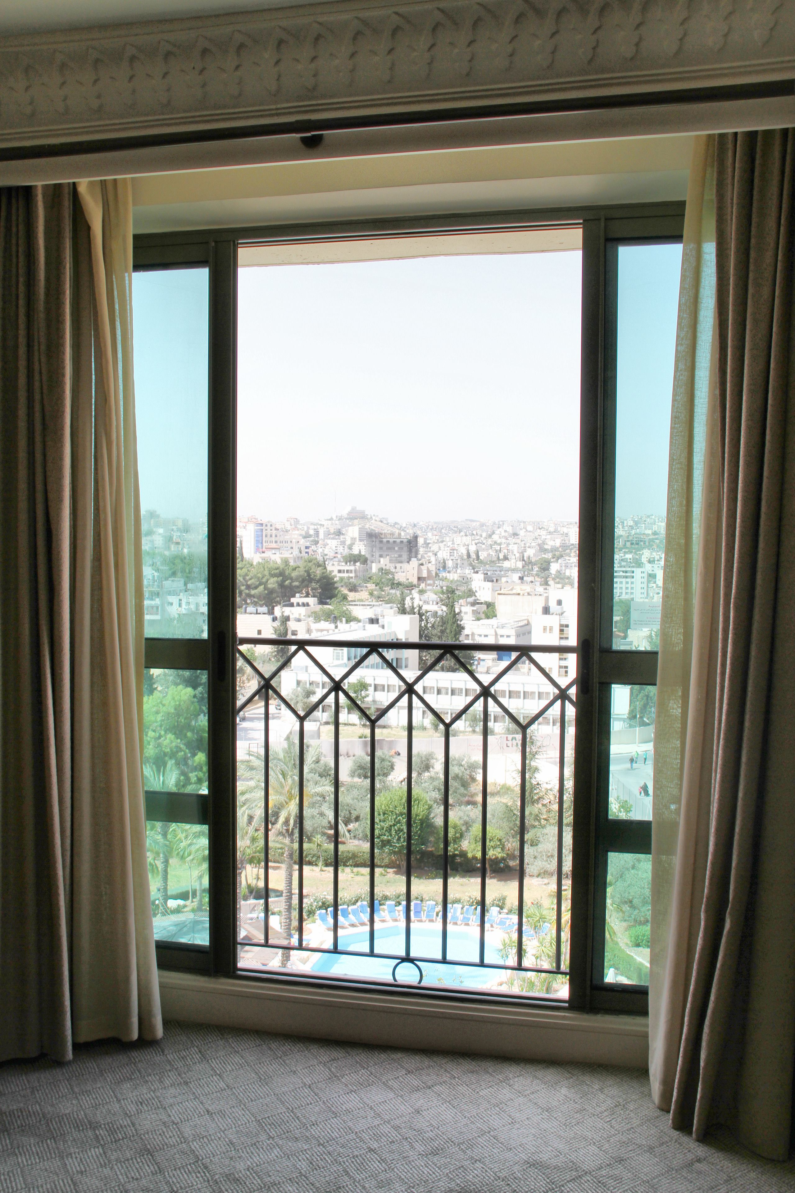 Una habitación con un amplio ventanal, cortinas y vistas a un paisaje urbano y una piscina.