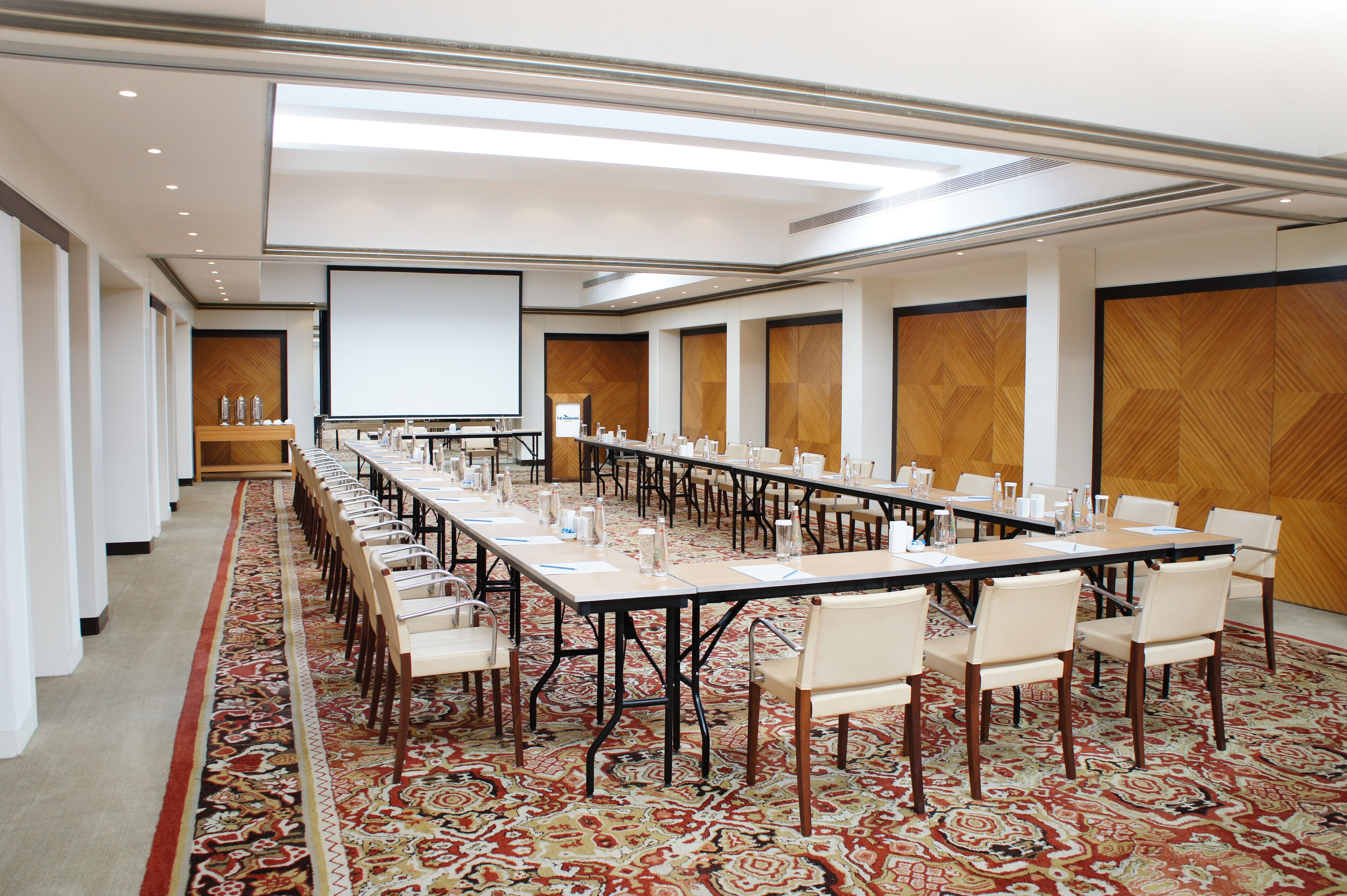 A conference room with a long table, chairs, a projector screen, and a patterned carpet.