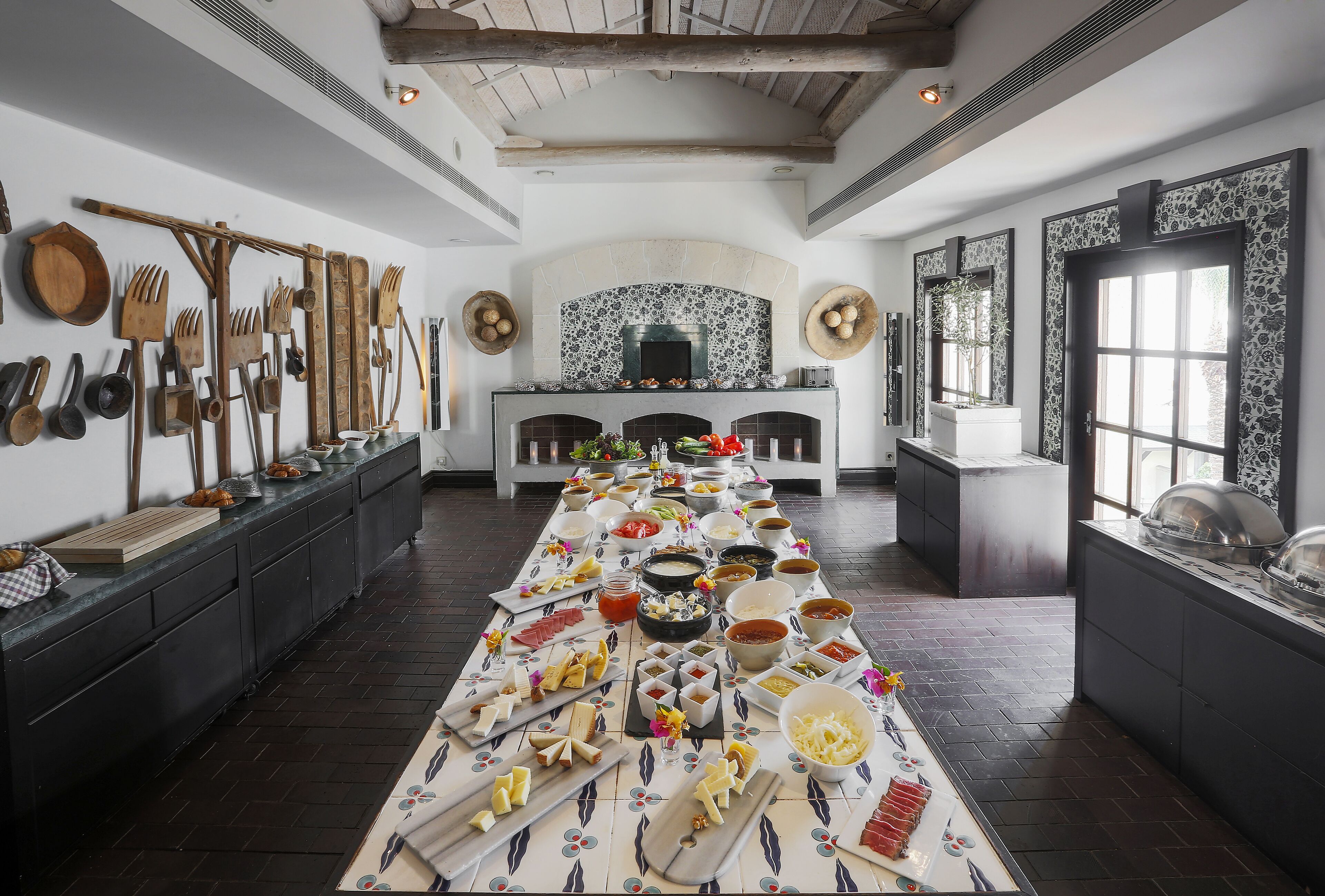 Uma longa mesa de jantar com diversos alimentos e utensílios, uma lareira e um teto rústico.