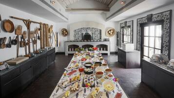 Uma longa mesa de jantar com diversos alimentos e utensílios, uma lareira e um teto rústico.