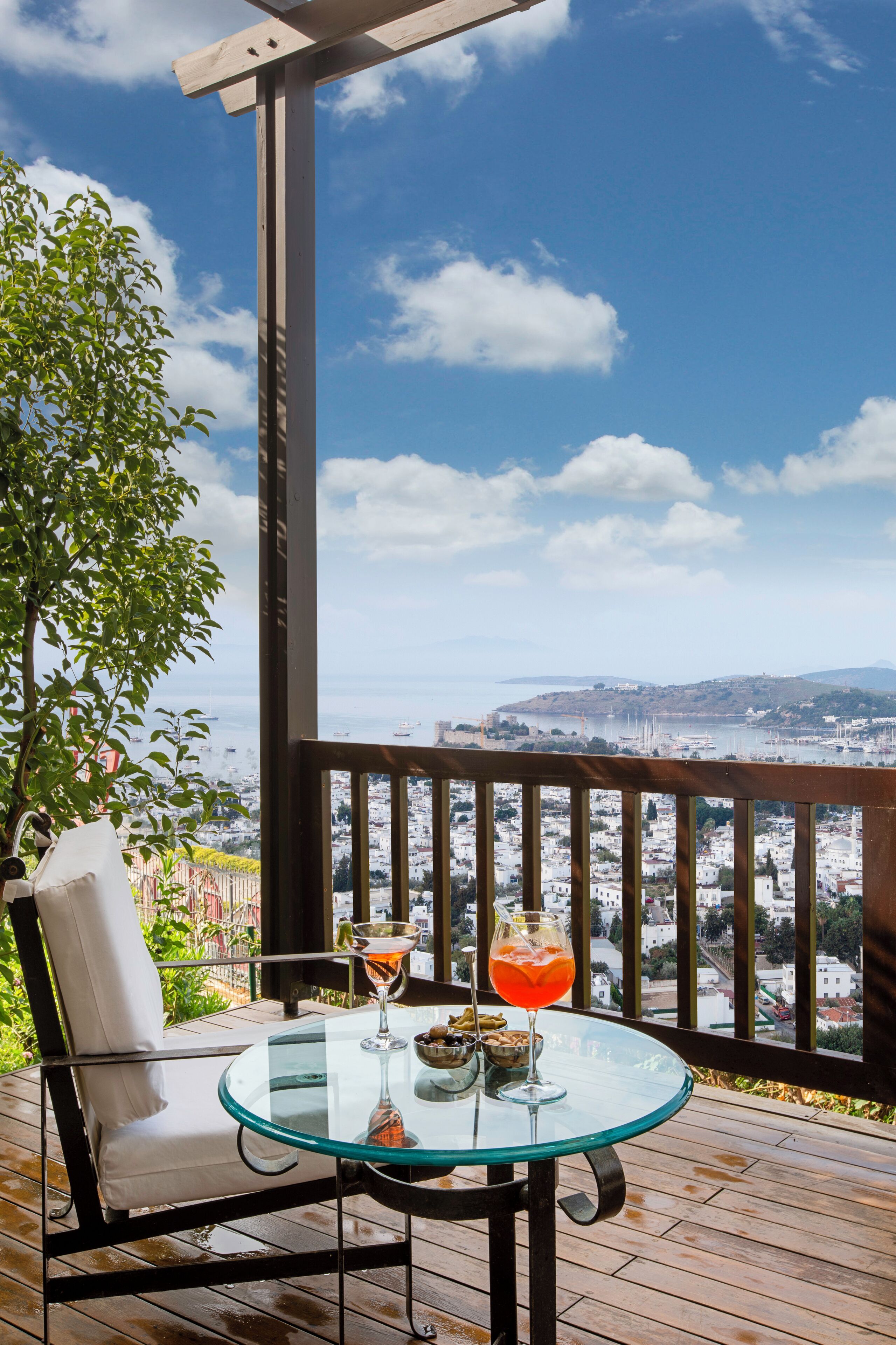 A glass table with two drinks and a bowl of nuts on a wooden deck with a view of a city and hills.