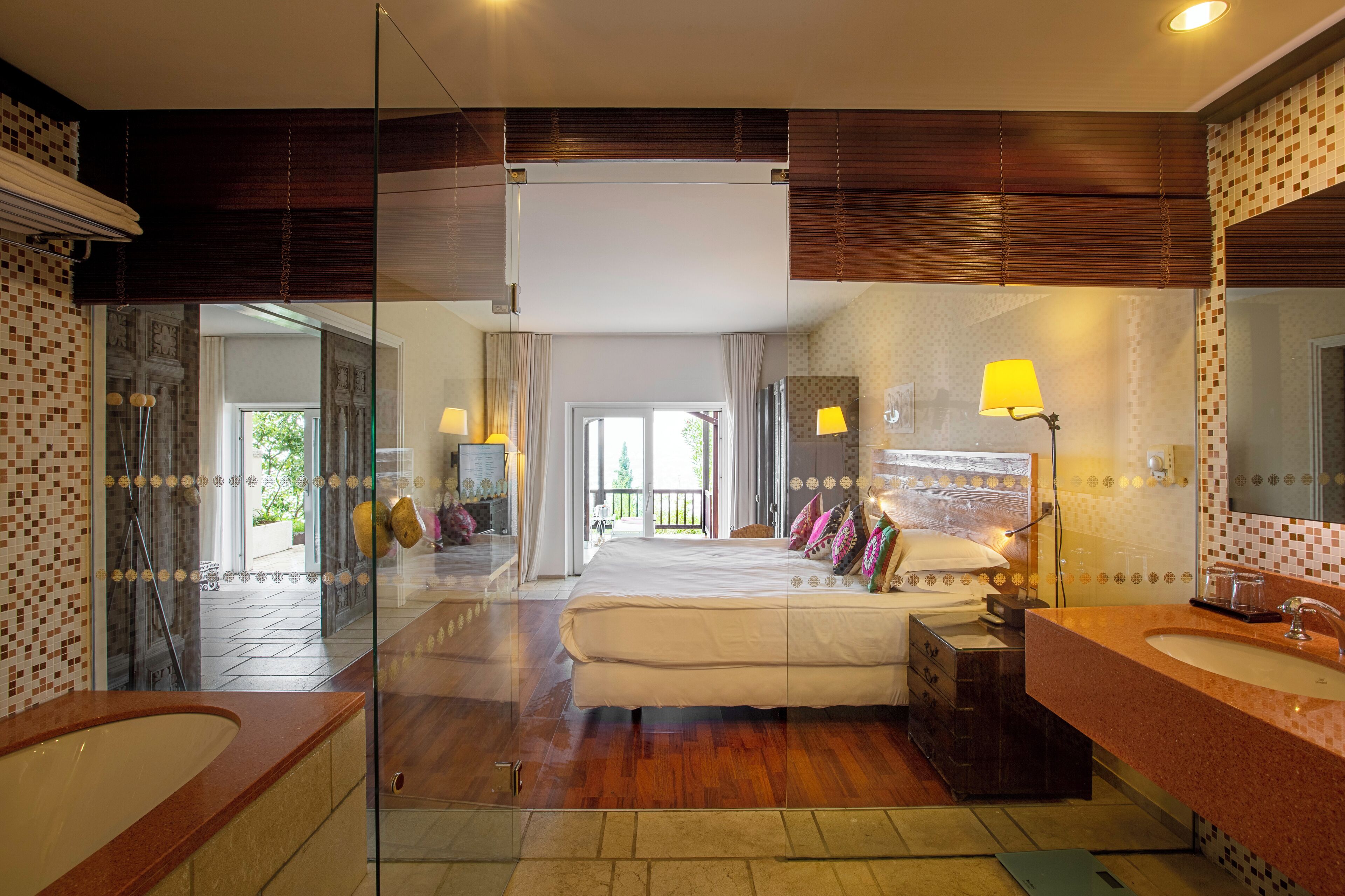 A modern hotel room with a large bed, a glass-enclosed shower, a separate bathroom with a sink, and a view of the outdoors.