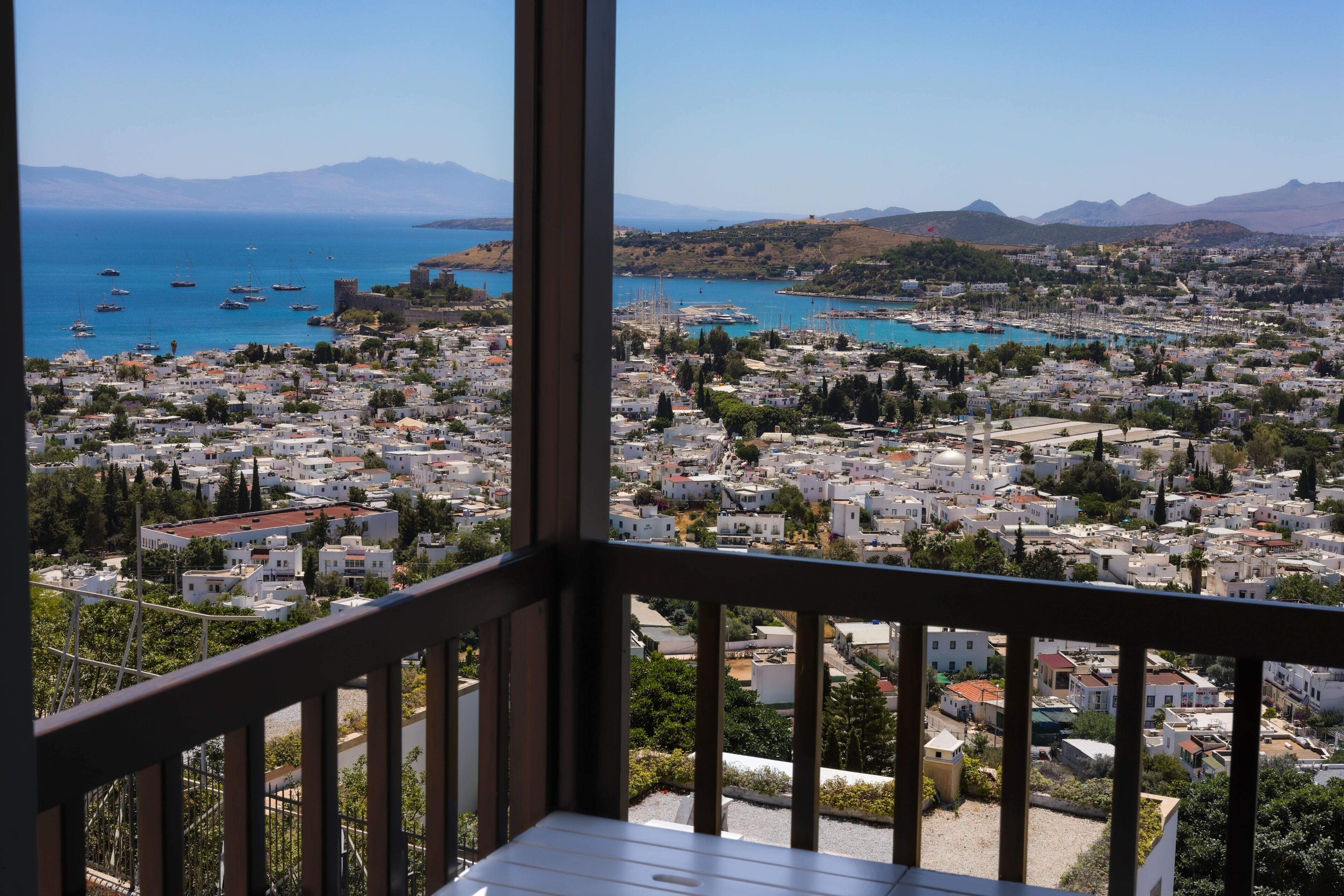 A balcony with a view of a coastal town, including buildings, boats, and mountains.
