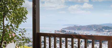 A glass table with two drinks and a bowl of nuts on a wooden deck with a view of a city and hills.
