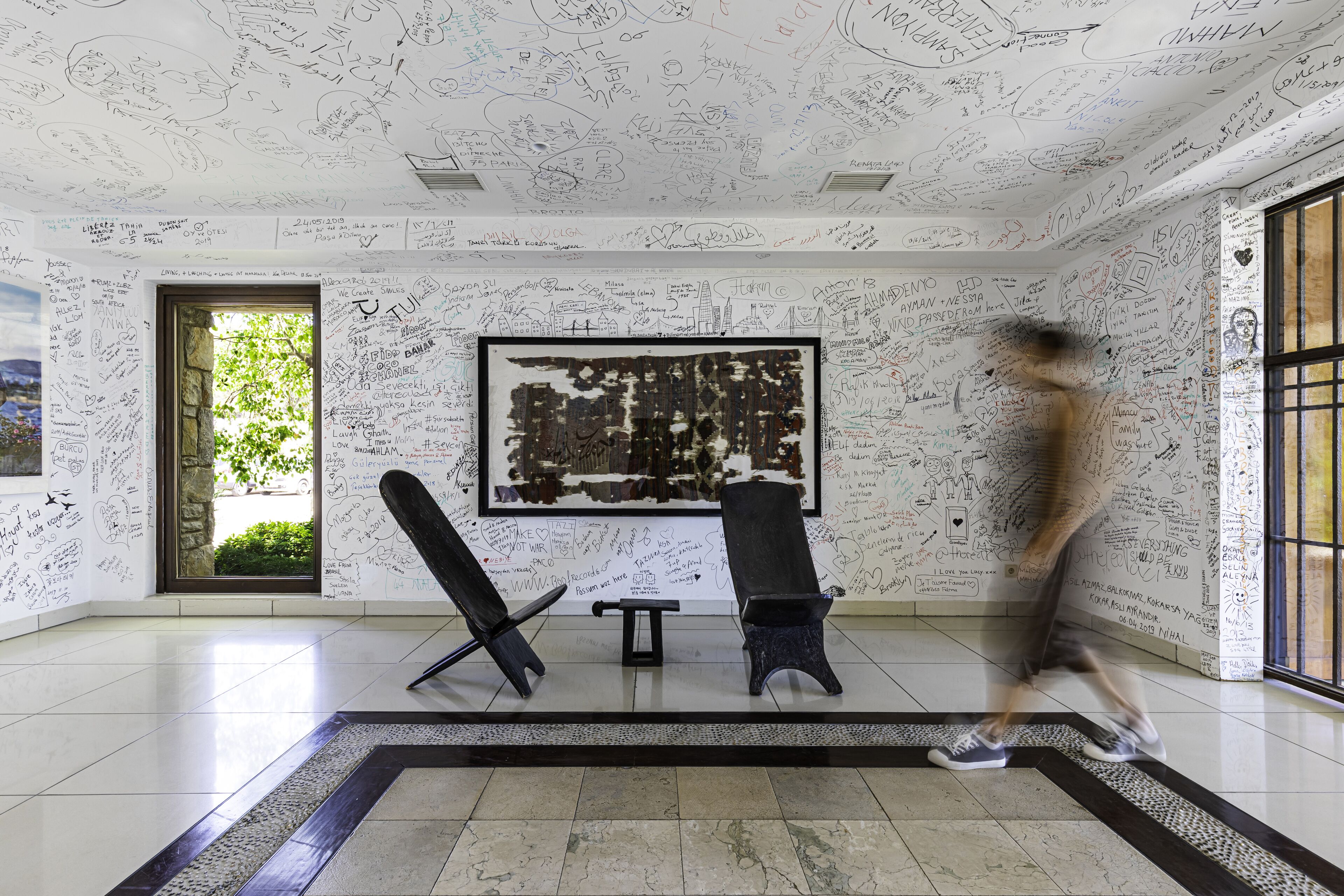 Une pièce aux murs blancs recouverts de divers écrits et dessins, un grand tableau abstrait accroché au mur, et deux chaises noires disposées de part et d’autre d’un banc placé au centre.