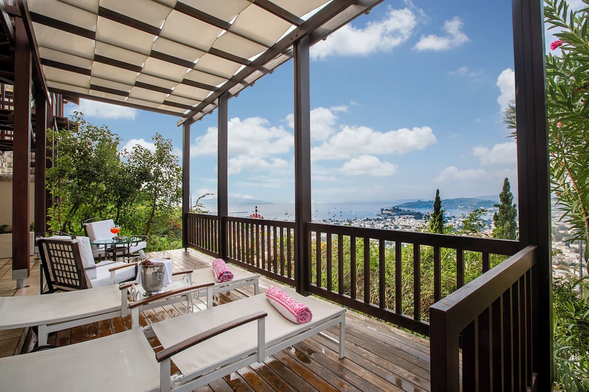 A wooden deck with white lounge chairs and a table, overlooking a scenic view of a bay with boats and hills.