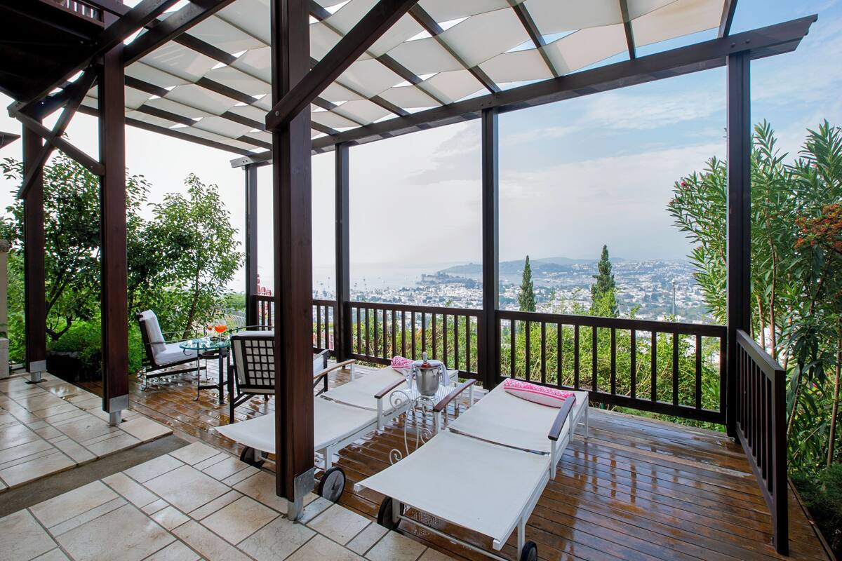 A covered outdoor area with a wooden deck, white lounge chairs, and a table with chairs.