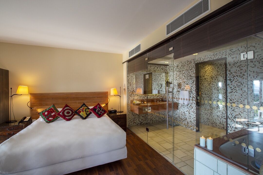 A hotel room with a bed, bedside lamps, a bathroom with a glass partition, and a mirror.