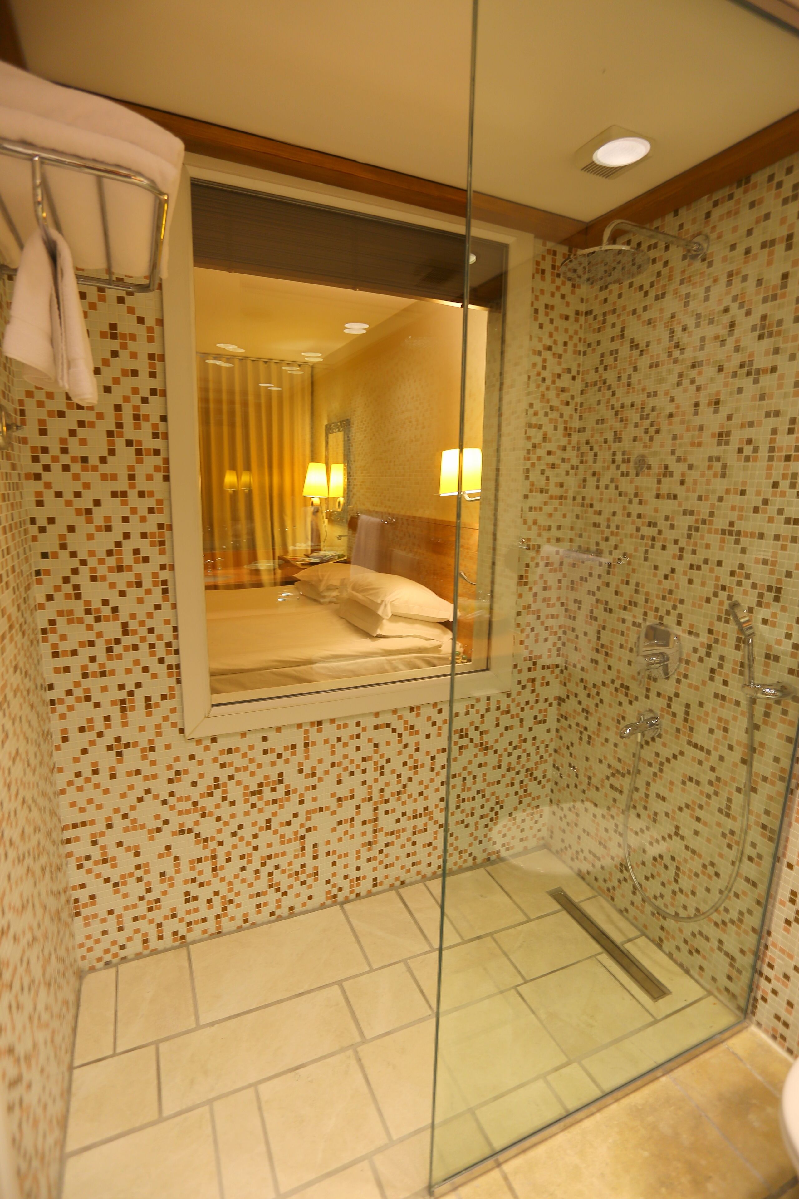 A glass-enclosed shower with mosaic tile walls, a visible bed with white linens, and a bathroom with a tiled floor.