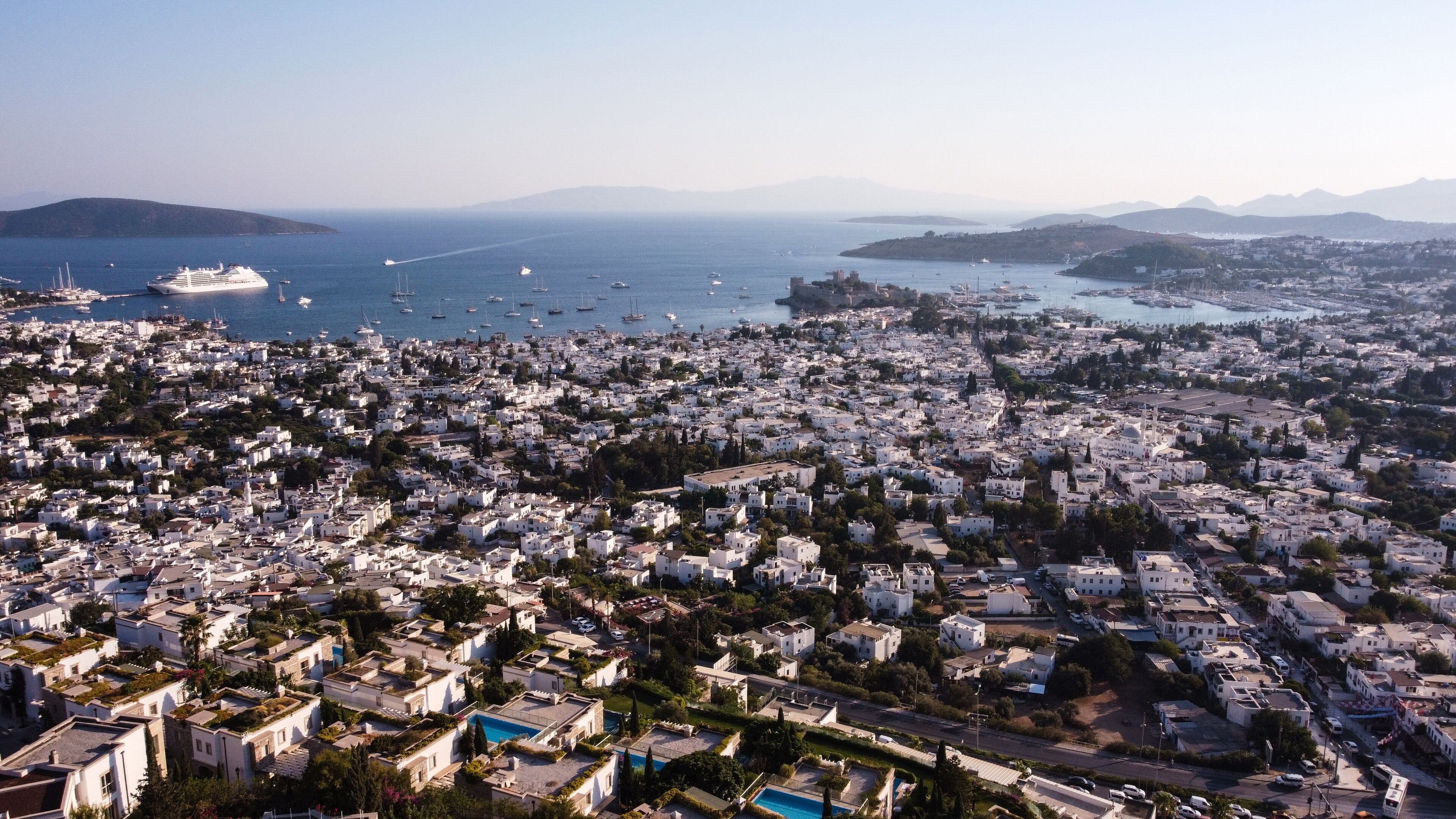 An aerial view of a coastal city with dense white buildings, a marina with boats, and a clear view of the sea and distant mountains.