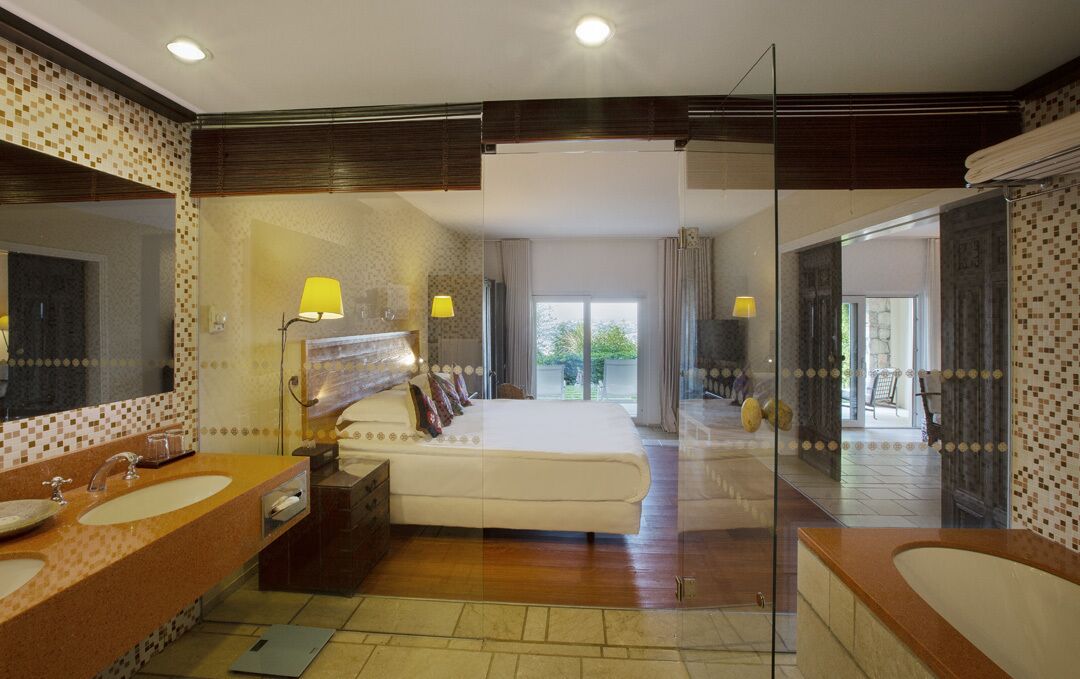 Habitación de hotel moderna con una cama grande, dos lavamanos, un balcón con paredes de vidrio y un espejo amplio.