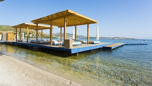 A waterfront resort with wooden cabanas, clear water, and a distant view of a hilly landscape. - The Marmara Bodrum - Adult Only (Bodrum)