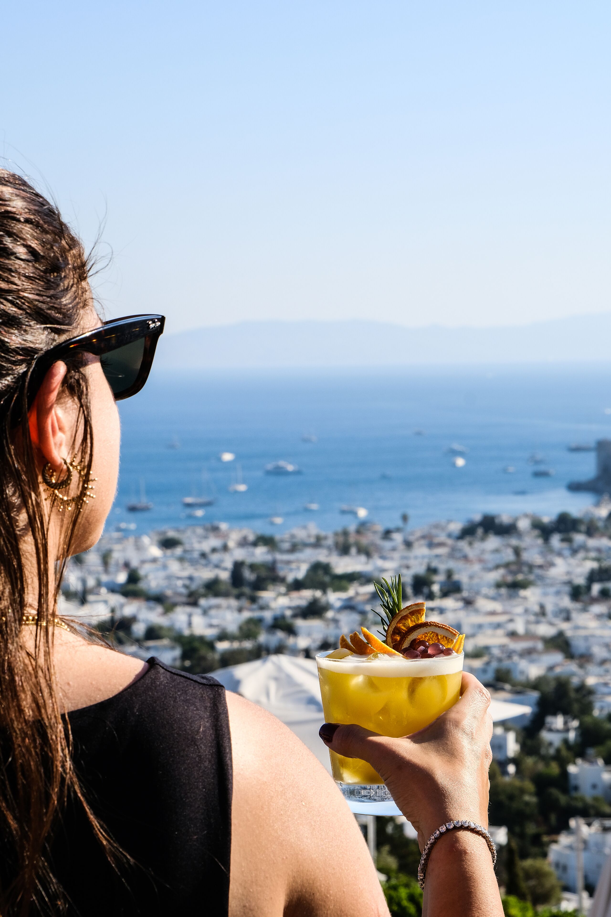 A person holding a cocktail with a view of the ocean and cityscape in the background.