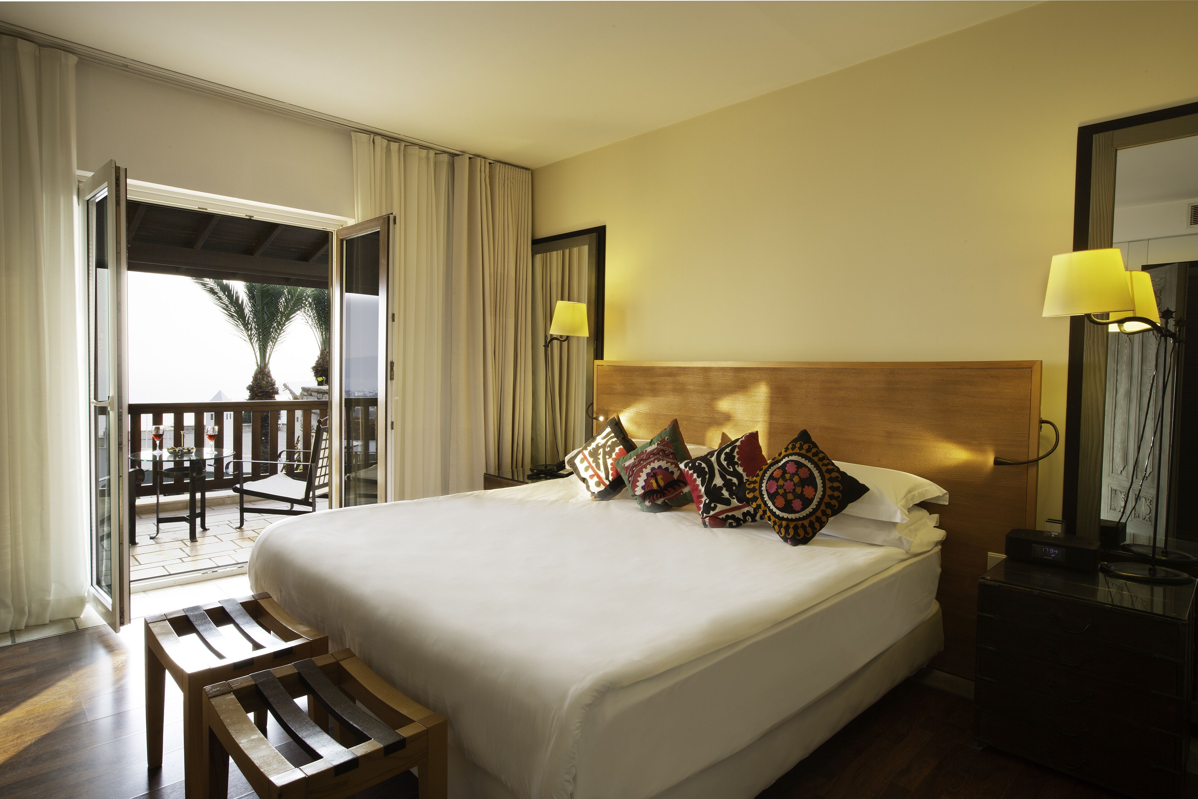 A hotel room with a large bed, a wooden bench, a balcony with a table and chairs, and two bedside lamps.
