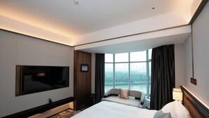Habitación de hotel con una cama grande, un televisor montado en la pared, un sofá y vistas a un paisaje urbano por la ventana.