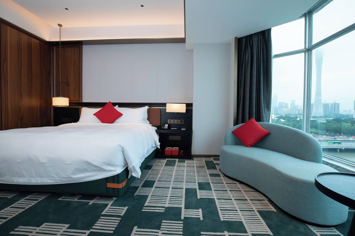 Hotelkamer met een groot bed, een bank, een kleine tafel en uitzicht op een stadslandschap door grote ramen.