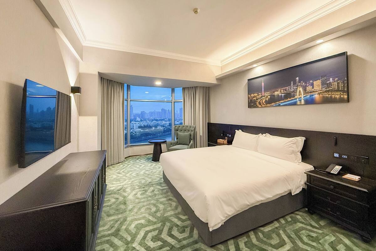 Een hotelkamer met een groot bed, een bureau, een stoel, een nachtkastje, een televisie en een raam met uitzicht op de stad.