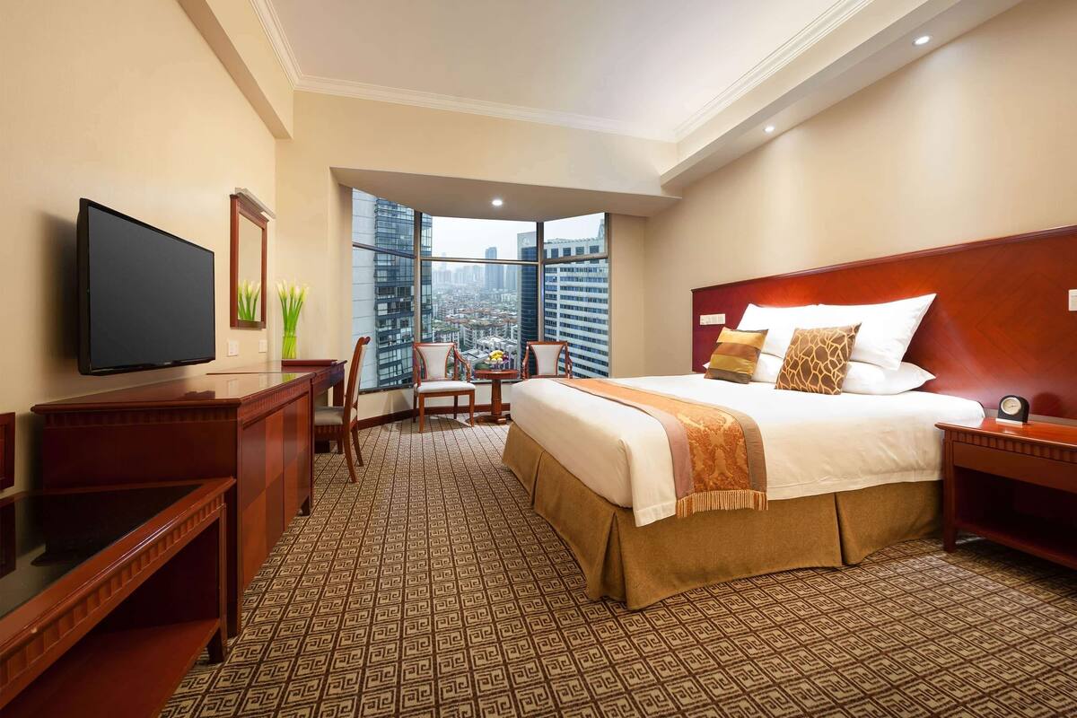 Hotelkamer met een groot bed, een bureau, een televisie en uitzicht op de stad door grote ramen.