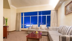 Habitación de hotel con un ventanal grande, un conjunto de sofá, una mesa de centro de madera y vistas a un paisaje urbano nocturno.