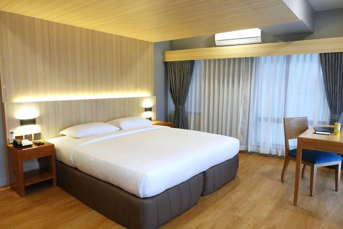 A hotel room with a large bed, bedside lamps, a desk with a chair, and a window with curtains.