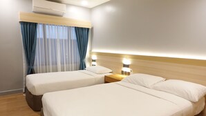 Habitación de hotel con dos camas, cabecero de madera, mesita de noche con lámpara y ventana con cortinas translúcidas.