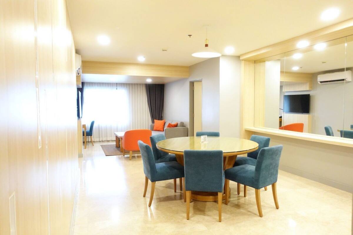 A modern hotel room with a round dining table, six blue chairs, a sofa with orange cushions, a TV, and an air conditioning unit.