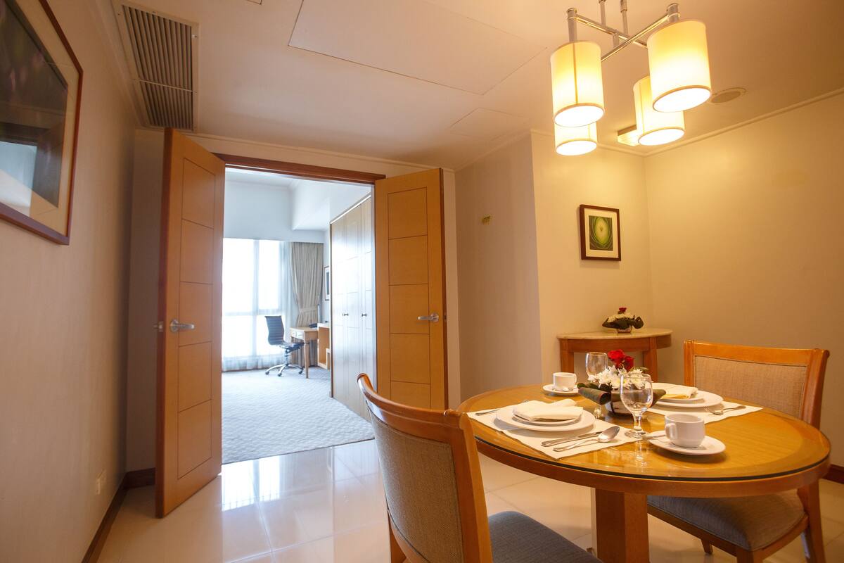A hotel dining area with a round table set for two, equipped with plates, cutlery, and a glass. There are two chairs, a small table with a vase of flowers, and a picture on the wall.