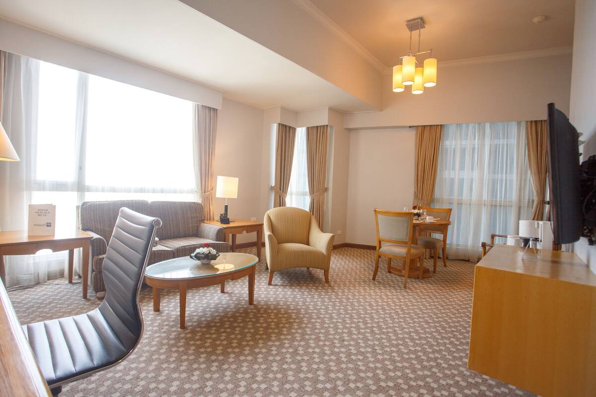 A hotel room with a sofa, armchair, dining table, and a TV.