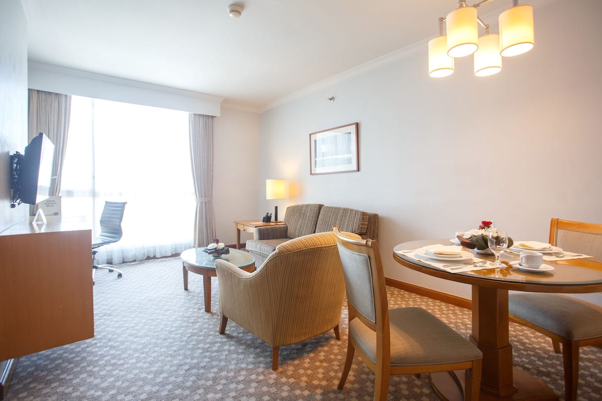 A hotel room with a dining table, chairs, a sofa, a TV, and a lamp.