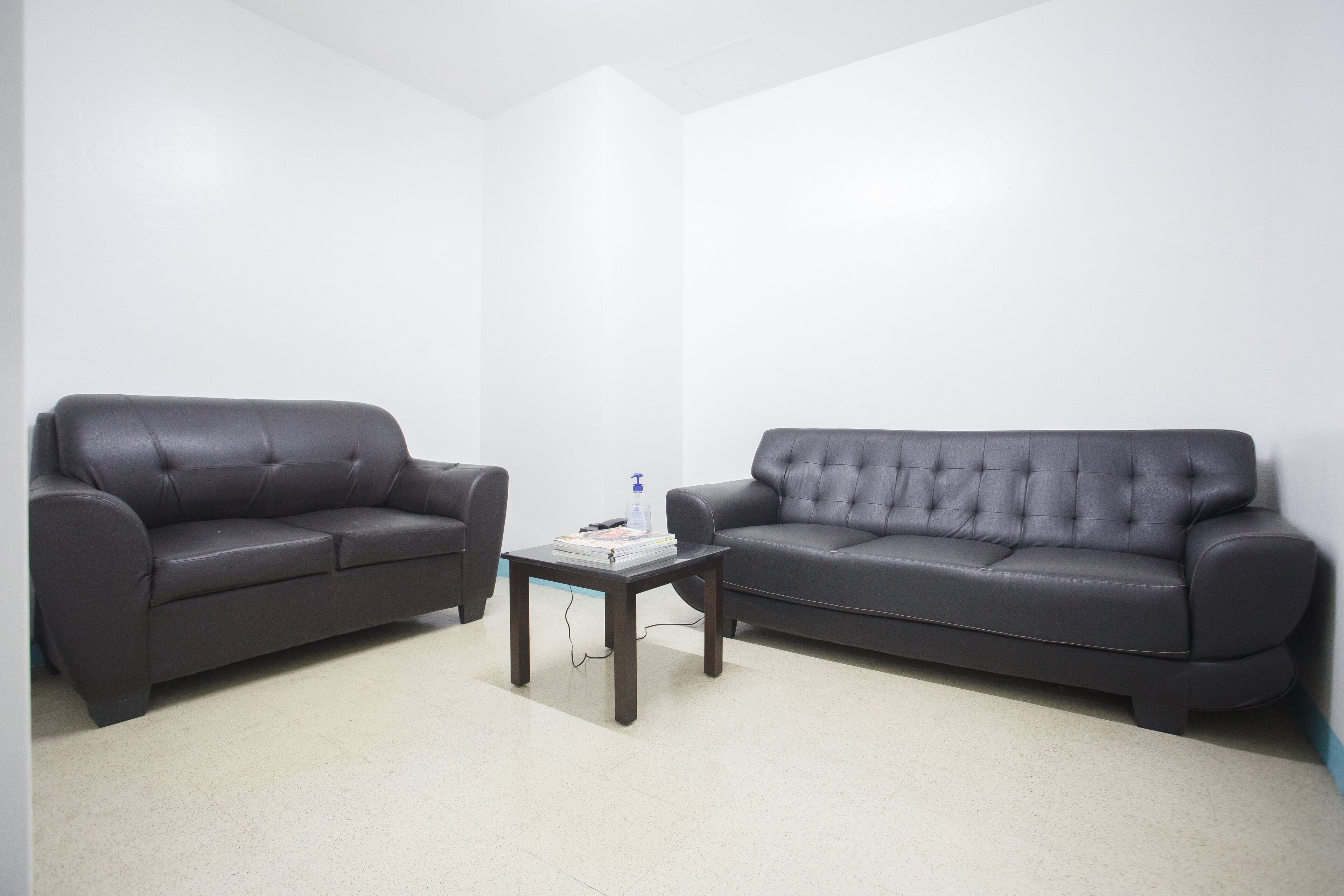 A room with two dark grey sofas, a small wooden coffee table with a book and a bottle, and a plain white wall.