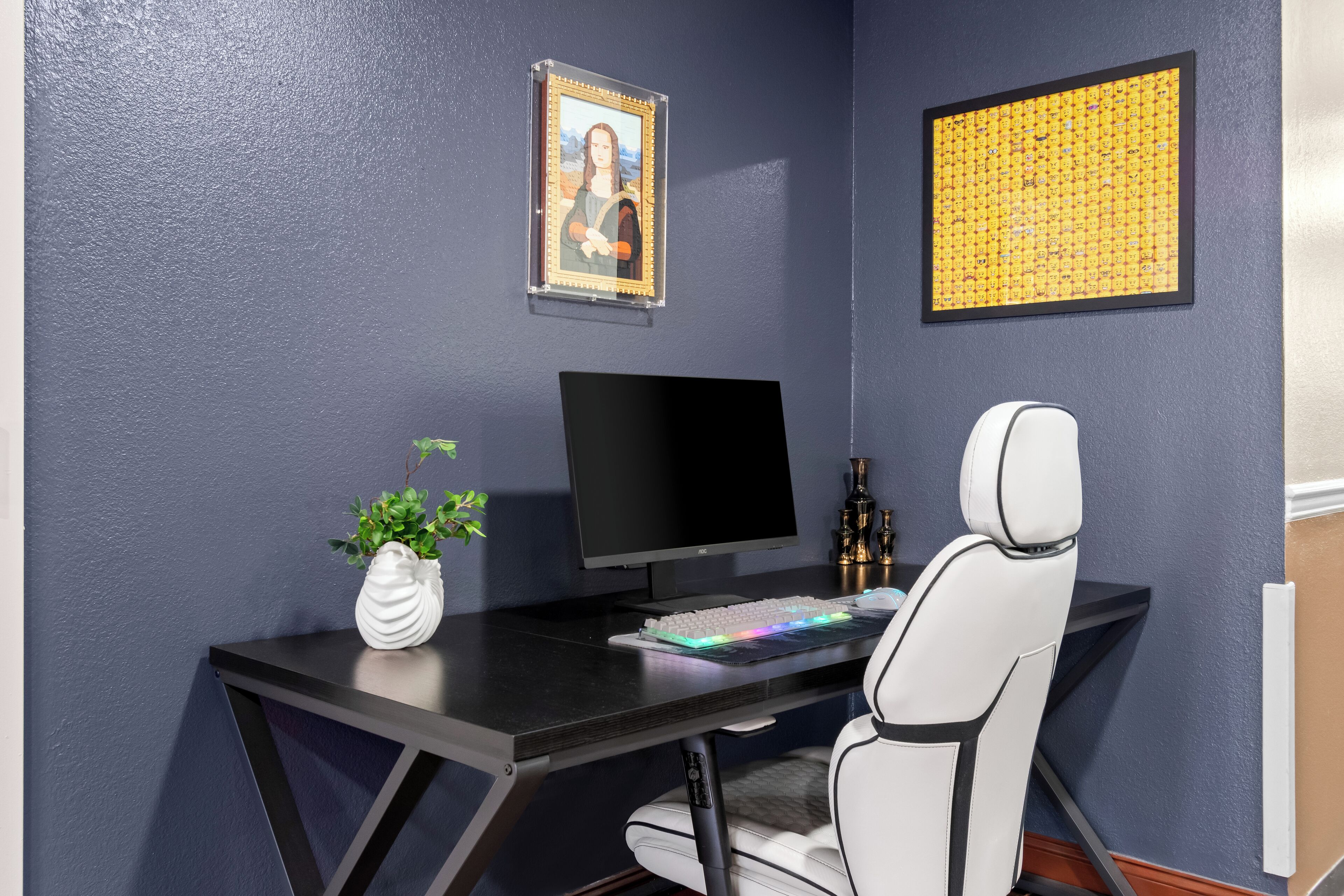 A modern office desk with a computer, keyboard, and mouse. There is a white chair, a small plant, and two framed pictures on the wall.