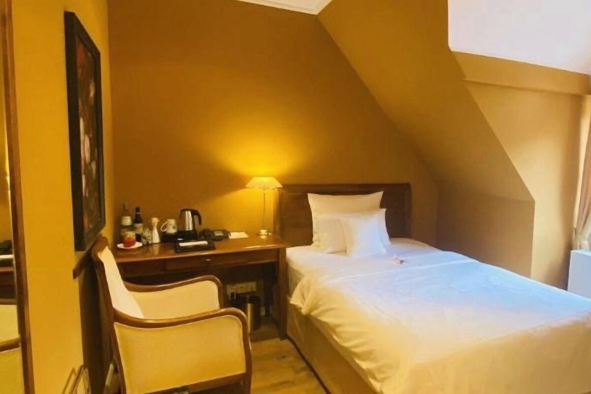 A hotel room with a bed, a desk, a chair, a lamp, and a window.