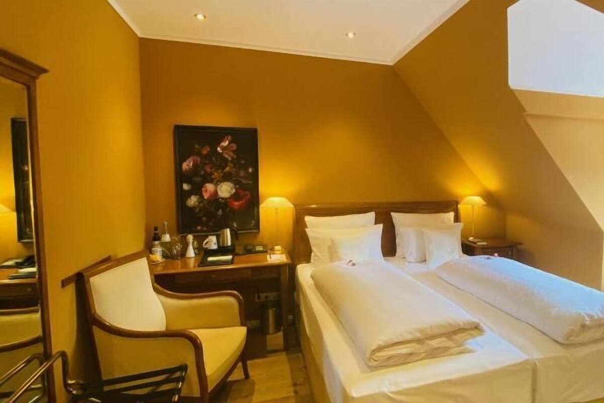 A hotel room with a bed, desk, chair, and a painting on the wall.