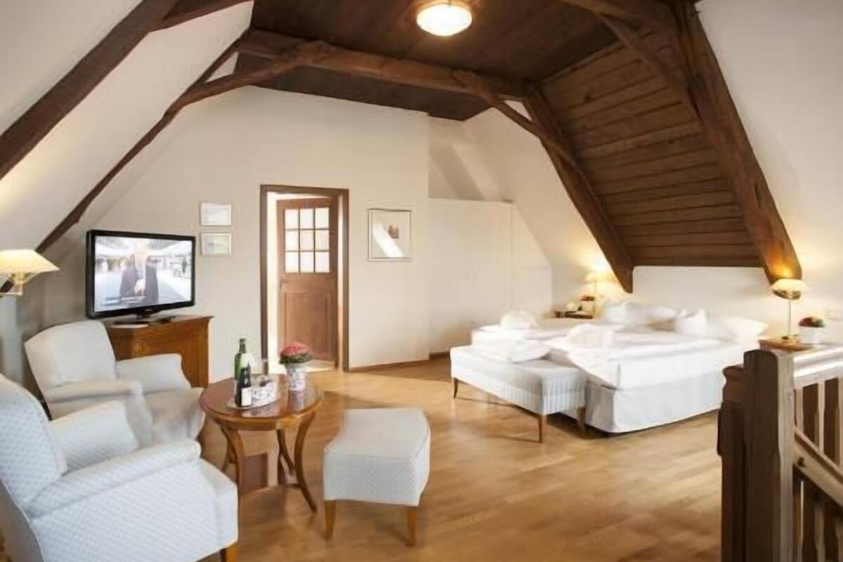 A cozy attic bedroom with a wooden ceiling, a television, a bed, and a small table with a lamp.