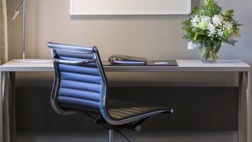 A modern office desk with a blue office chair, a white desk lamp, a framed abstract art piece, and a vase with flowers.