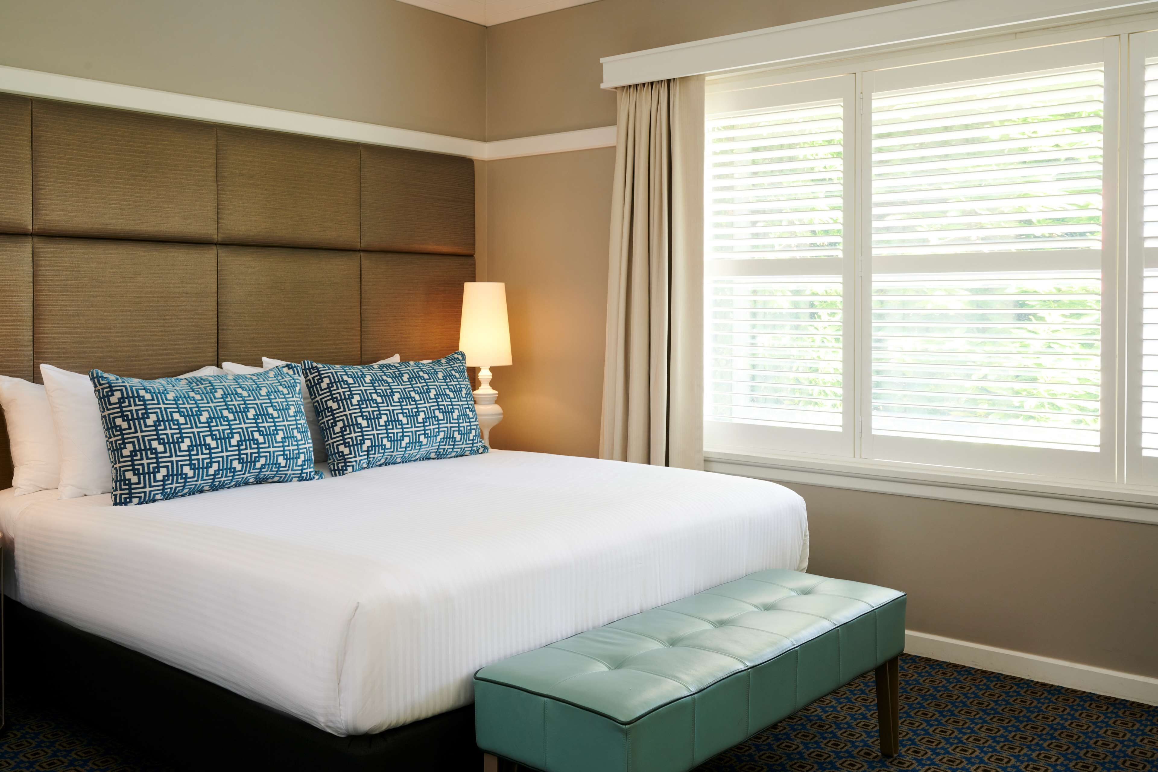 A hotel room with a large bed, a headboard, a window with blinds, a bedside lamp, and a bench.