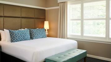A hotel room with a large bed, a headboard, a window with blinds, a bedside lamp, and a bench.