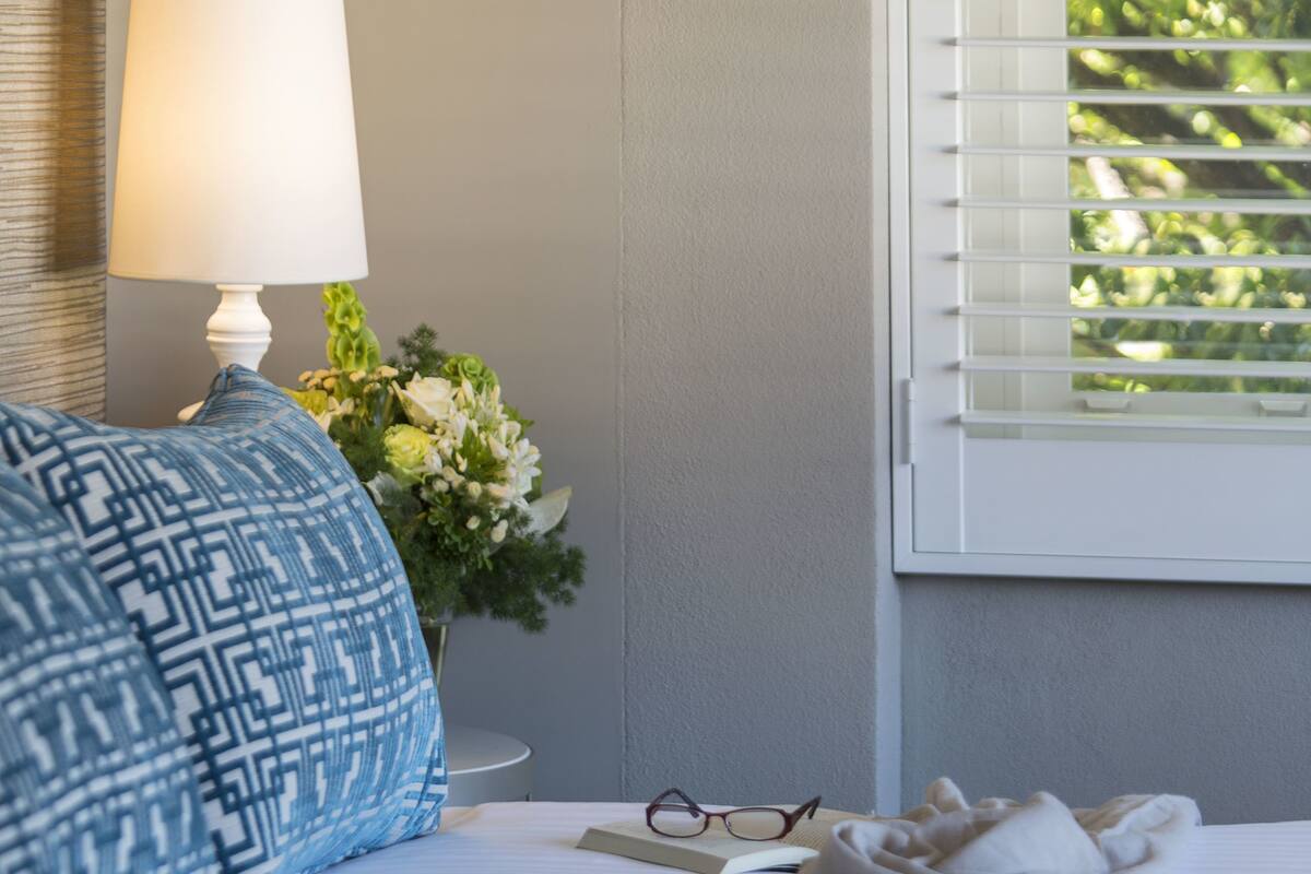 A neatly made bed with a white comforter, a blue patterned pillow, a white lamp, and a vase with flowers on a bedside table.