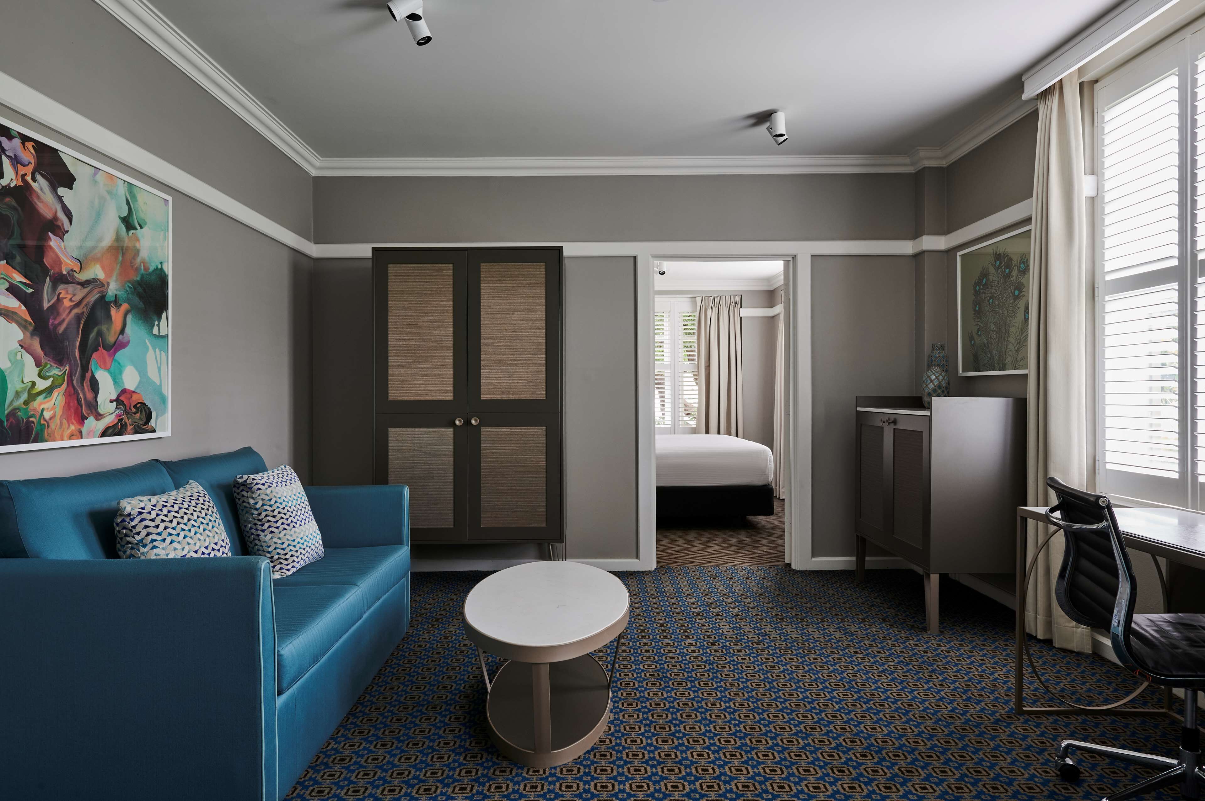 Habitación de hotel con sofá azul, una mesa redonda, un escritorio con silla, armario, cama y un cuadro abstracto grande en la pared.