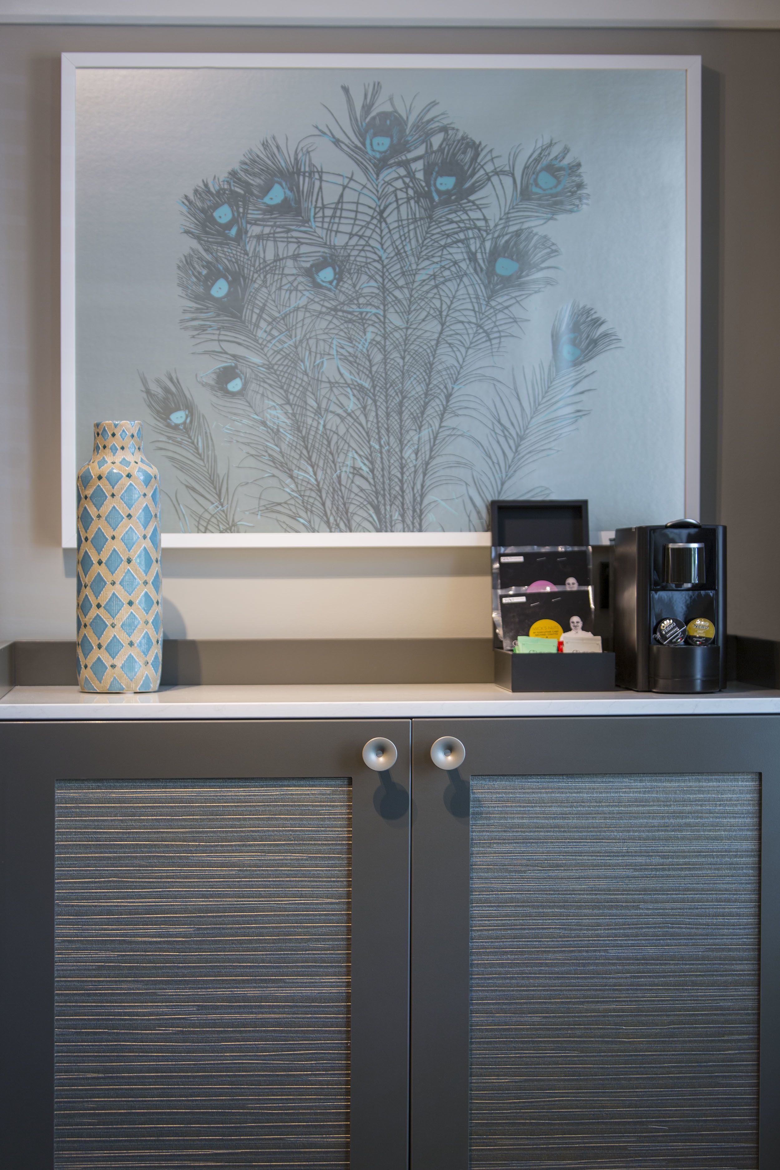 A framed artwork of peacock feathers, a decorative vase, and a coffee machine on a cabinet.